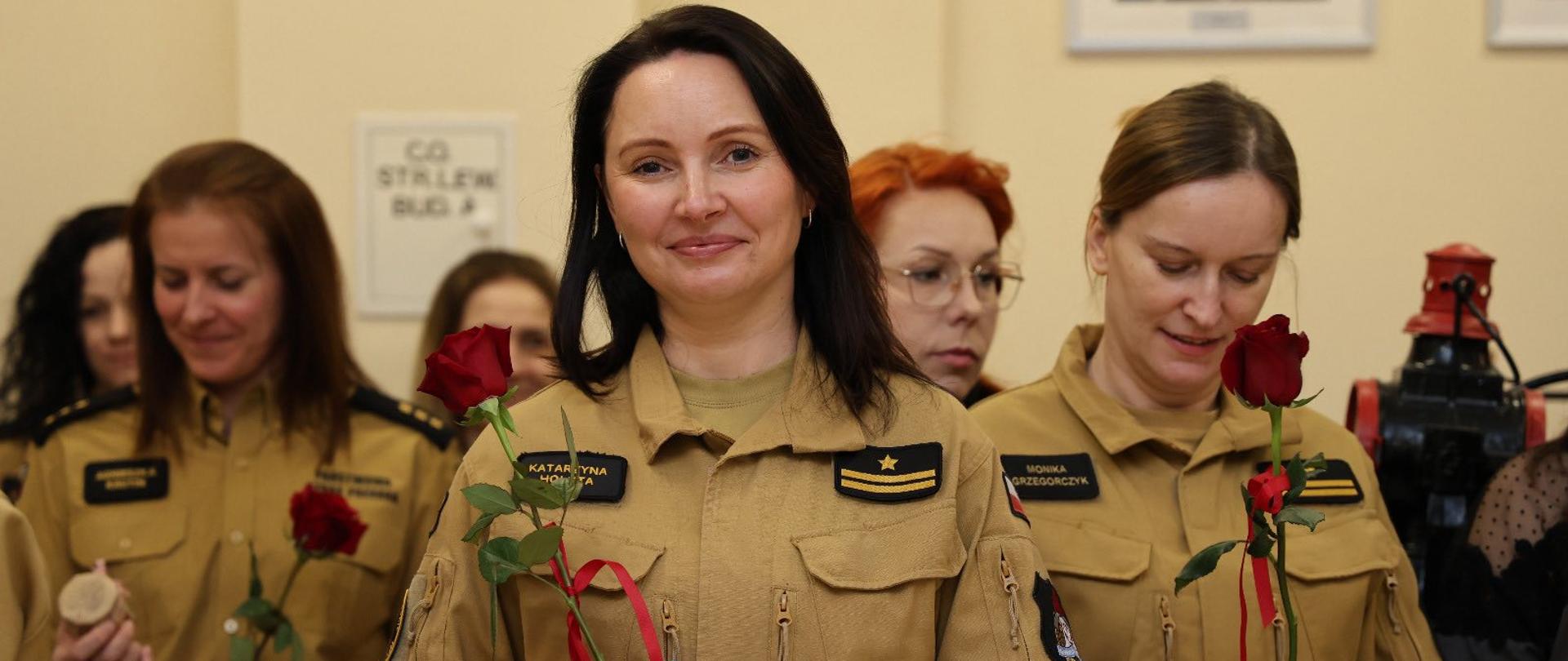 Grupa kobiet w mundurach strażackich stoi w pomieszczeniu podczas uroczystości. W rękach trzymają czerwone róże przewiązane wstążką oraz małe pudełka z upominkami. Na pierwszym planie uśmiechnięta strażaczka patrzy w stronę aparatu. Pozostałe osoby stoją w tle w podobnych mundurach.