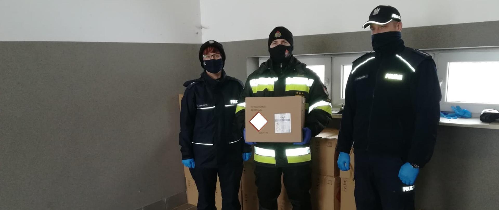 Zdjęcie przedstawia jednego strażaka trzymającego karton ze środkami do dezynfekcji oraz dwóch funkcjonariuszy Policji (kobieta i mężczyzna). W tle większa ilość przekazanych środków do dezynfekcji w kartonach