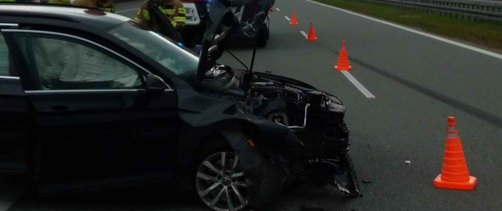 Zdjęcie przedstawia drogę ekspresową S7. Po lewej stronie stoi uszkodzony samochód osobowy. Za nim widać ratowników i samochód pożarniczy.