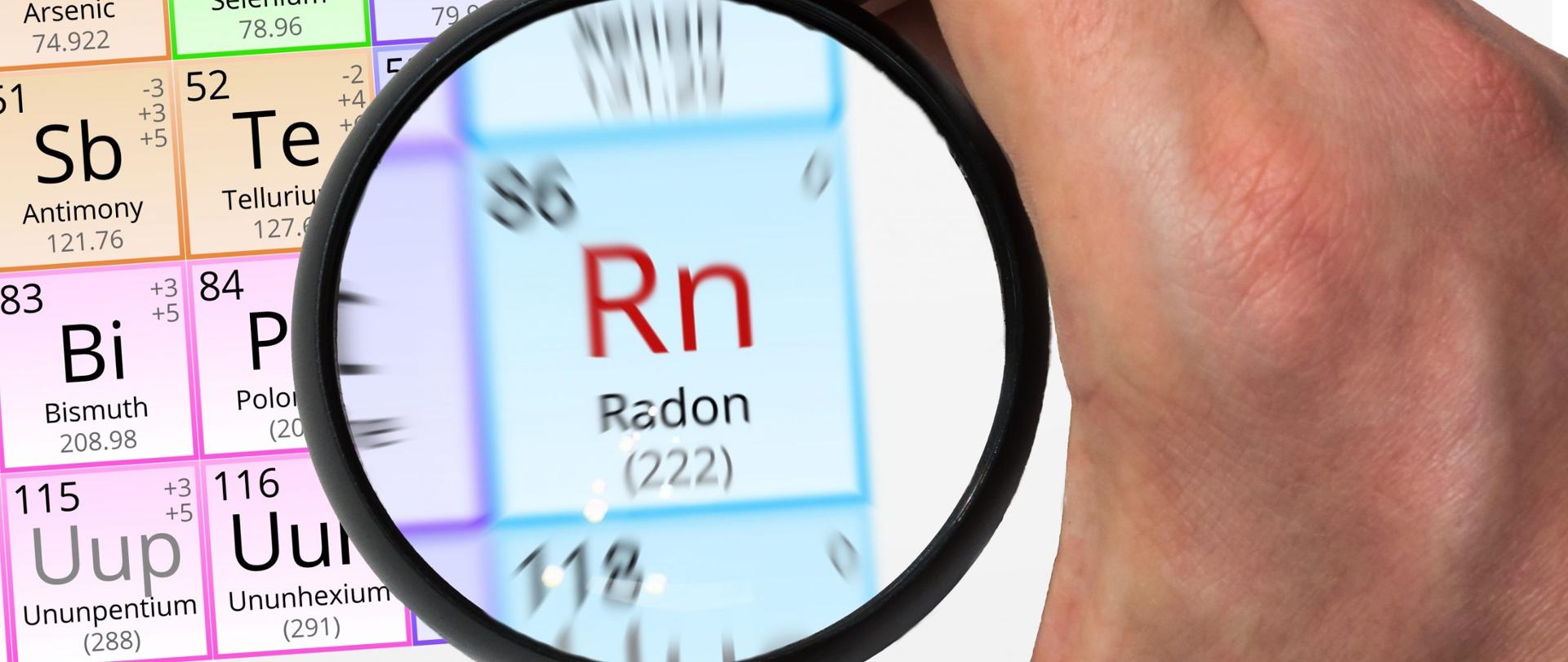 Na zdjęciu w tle widoczna jest tablica z pierwiastkami. Na pierwszym planie znajduje się dłoń trzymająca lupę skierowaną na pole Radonu.