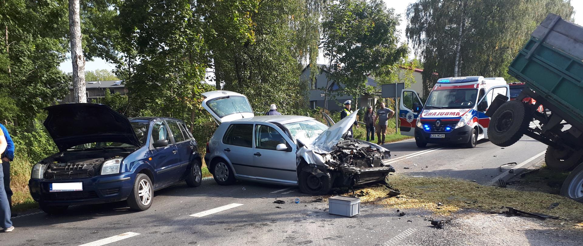 Zdjęcie przedstawia sytuację na drodze po wypadku z udziałem ciągnika rolniczego z przyczepą. Na pierwszym planie dwa pojazdy osobowe. W prawej części zdjęcia widoczna przewrócona przyczepa. W oddali ambulans.