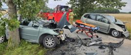 Na zdjęciu dwa mocno uszkodzone pojazdy osobowe blokujące ruch na drodze powiatowej. W tle parawan ochrony oraz pojazdy pożarnicze.