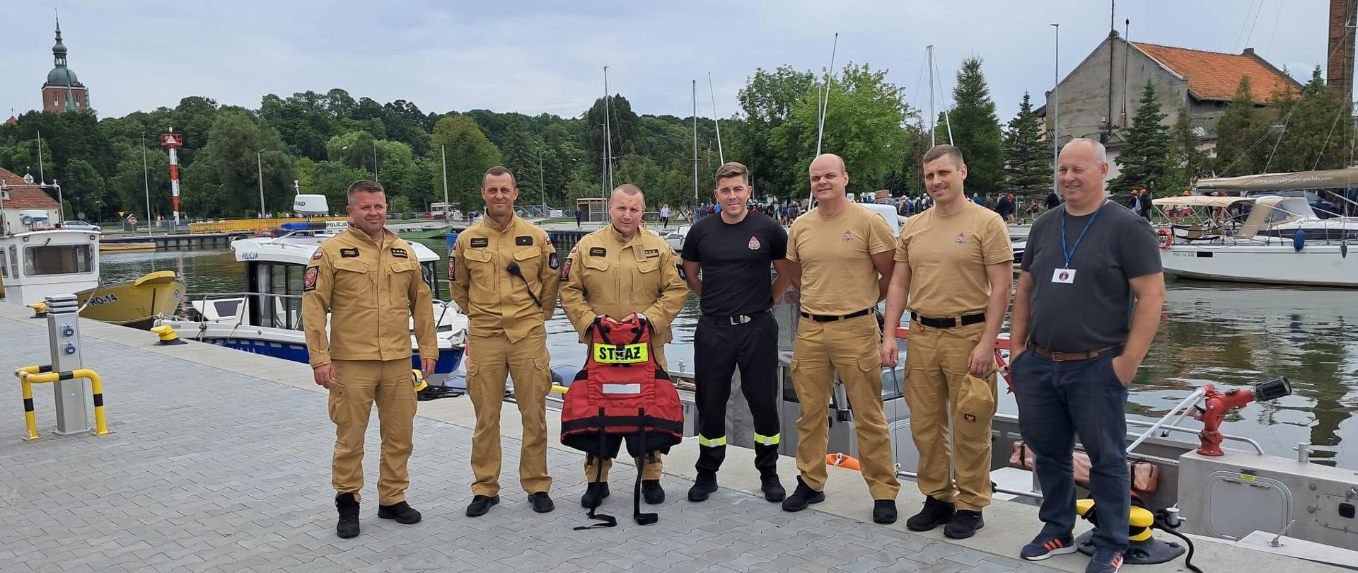 W porcie na brzegu stoi grupa strażaków z bosmanem. W tle w porcie stoją łodzie.