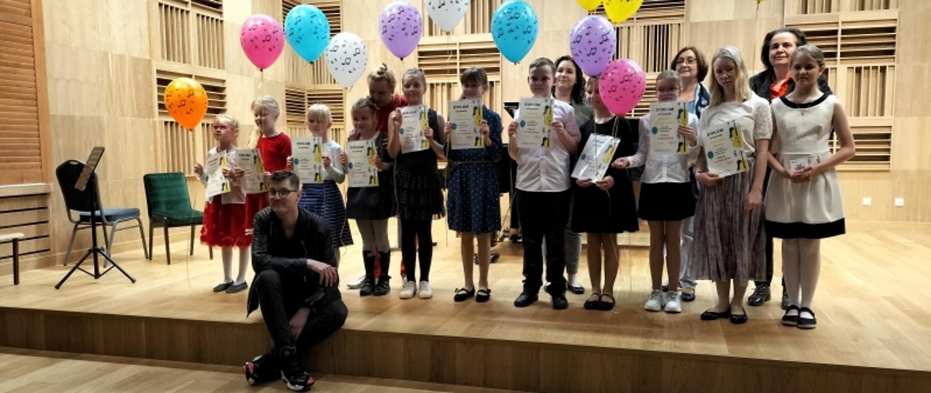 Grupa jedenastu uczniów klas pierwszych z dyplomami i balonikami w towarzystwie swoich nauczycieli na estradzie sali koncertowej Zespołu Szkół Muzycznych im. Fryderyka Chopina w Pile.