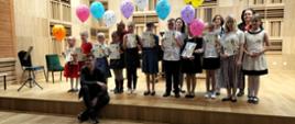 Grupa jedenastu uczniów klas pierwszych z dyplomami i balonikami w towarzystwie swoich nauczycieli na estradzie sali koncertowej Zespołu Szkół Muzycznych im. Fryderyka Chopina w Pile.