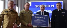 Trzech strażaków stoi z mężczyznom w garniturze. Strażak oraz mężczyzna trzymają bon na dwieście tysięcy złotych dla Specjalistycznej Grupy Poszukiwawczo-Ratowniczej Gdańsk. 