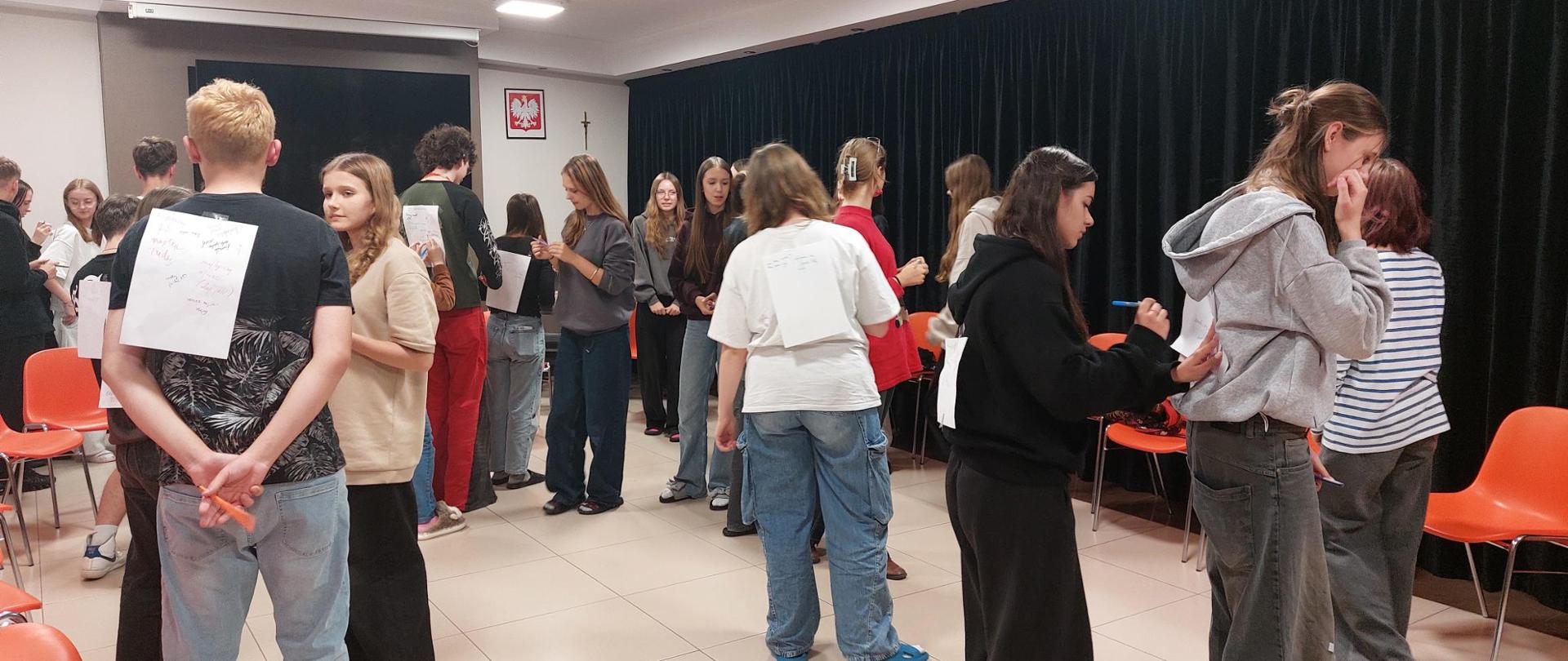 Około 20 osobowa grupa wychowanków znajduje się w dużej świetlicy. Stoją w rozproszeniu, na osobach stojących tyłem widać przyklejone kartki białego papieru na plecach. kilka osób coś na nich pisze