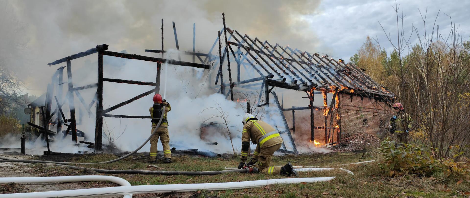 Na zdjęciu widoczny jest szkielet budynku gospodarczego, który gaszą strażacy. Widoczne są także płomienie oraz węże strażackie.