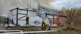 Na zdjęciu widoczny jest szkielet budynku gospodarczego, który gaszą strażacy. Widoczne są także płomienie oraz węże strażackie.