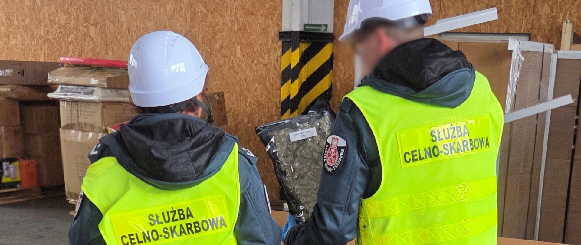 Dwie osoby w kamizelkach i kaskach ochronnych oglądają duży, przezroczysty worek z suszem roślinnym w hali magazynowej; wokół widoczne są kartony i materiały składowane na paletach.