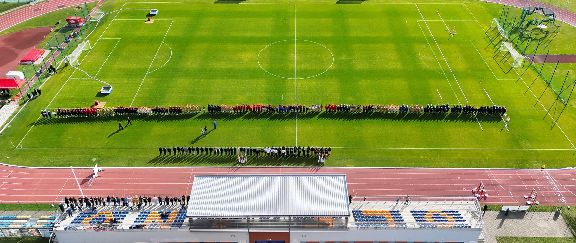 Widok z lotu ptaka na stadion miejski w Oleśnie podczas zawodów sportowo-pożarniczych. Na płycie boiska ustawieni są strażacy w umundurowaniu sportowym, a także sędziowie, zaproszeni goście i organizatorzy zawodów. Na stadionie widoczne są boisko piłkarskie, bieżnia lekkoatletyczna oraz urządzenia wykorzystywane podczas konkurencji. W dolnej części zdjęcia znajduje się trybuna z zadaszeniem i miejscami dla publiczności.