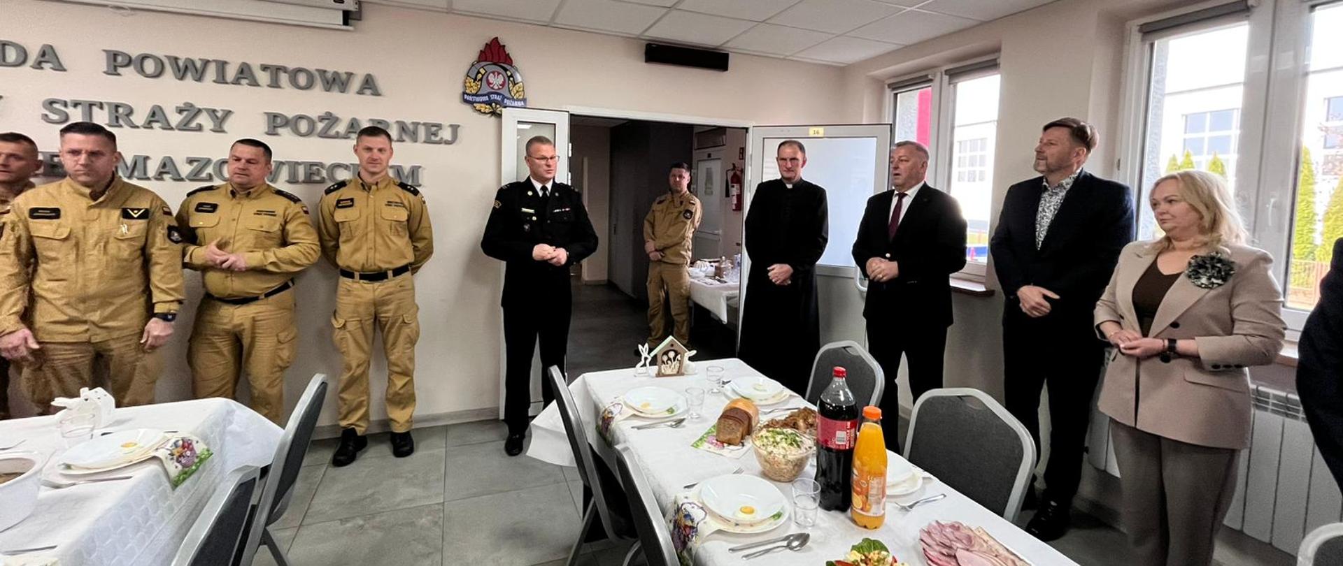 Zastępca Komendanta Powiatowego wita funkcjonariuszy, pracowników i przybyłych gości na spotkanie wielkanocne. Z prawej strony stoją zaproszeni goście, z lewej strony stoją funkcjonariusze PSP. 