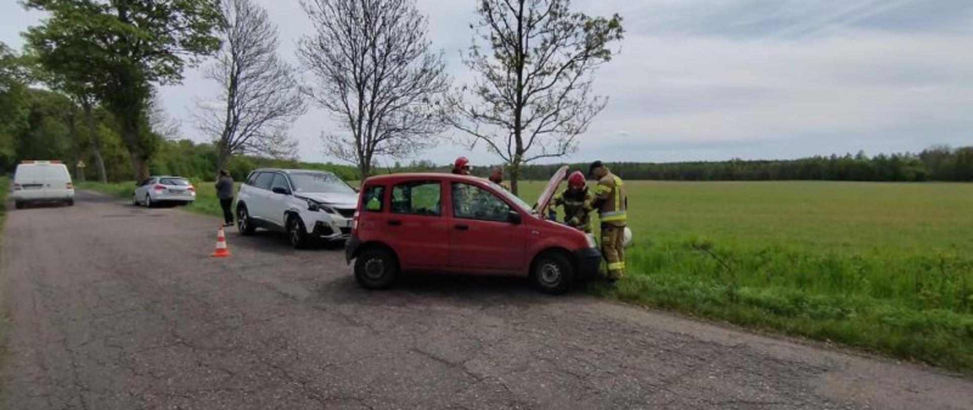 Zdjęcie wykonane w trakcie działań. Strażacy stoją przy czerwonym Fiacie Panda. Maska w aucie jest uniesiona a strażacy odłączają akumulator. W oddali drugi pojazd uczestniczący w wypadku praz pojazdy służb.