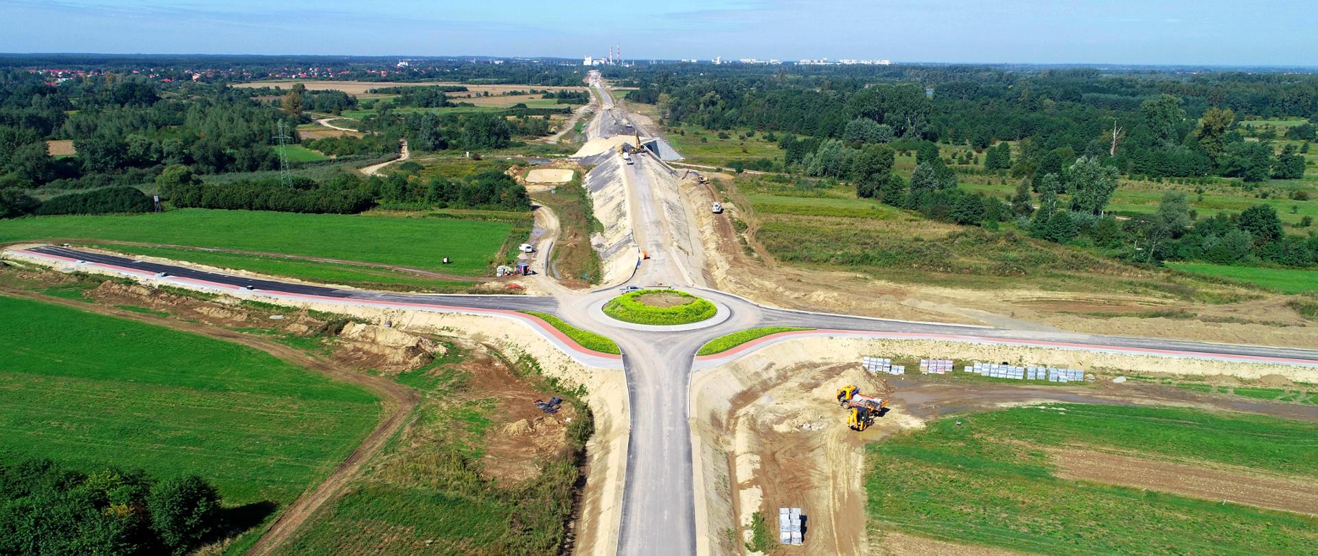 Na zdjęciu widać budowaną drogę jednojezdniową, po jednym pasie ruchu w każdą stronę. Pobocza są zarośnięte trawą w kolorze zielonym, w oddali widać wysokie, zielone drzewa.