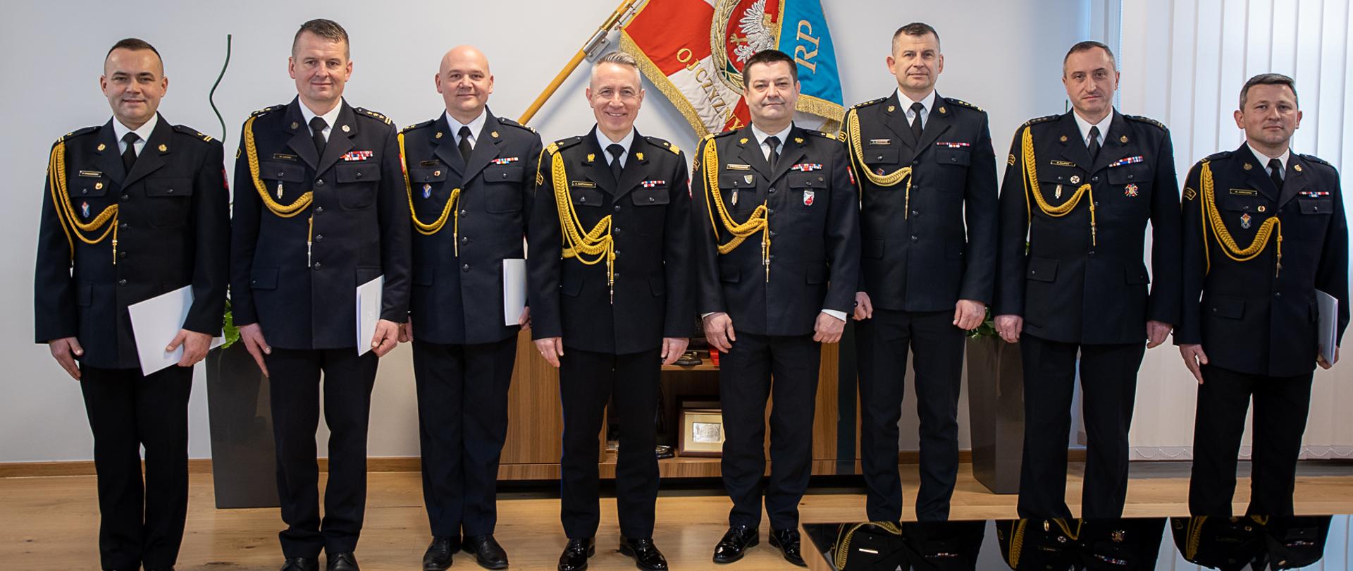 Na zdjęciu widać ośmiu strażaków w umundurowaniu wyjściowym. W tle sztandar państwowej Straży Pożarnej