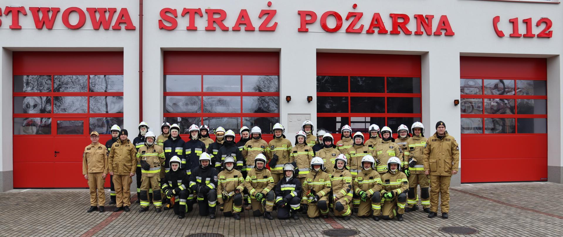 Na zdjęciu znajduje się duża grupa strażaków, w tym kilkudziesięciu dorosłych i kilku młodszych osób, wszyscy w mundurach strażackich i hełmach, stoi i klęczy przed budynkiem Komendy Powiatowej PSP w Przysusze. Budynek ma czerwone drzwi garażowe i napis "PAŃSTWOWA STRAŻ POŻARNA" oraz numer "112" nad nimi.