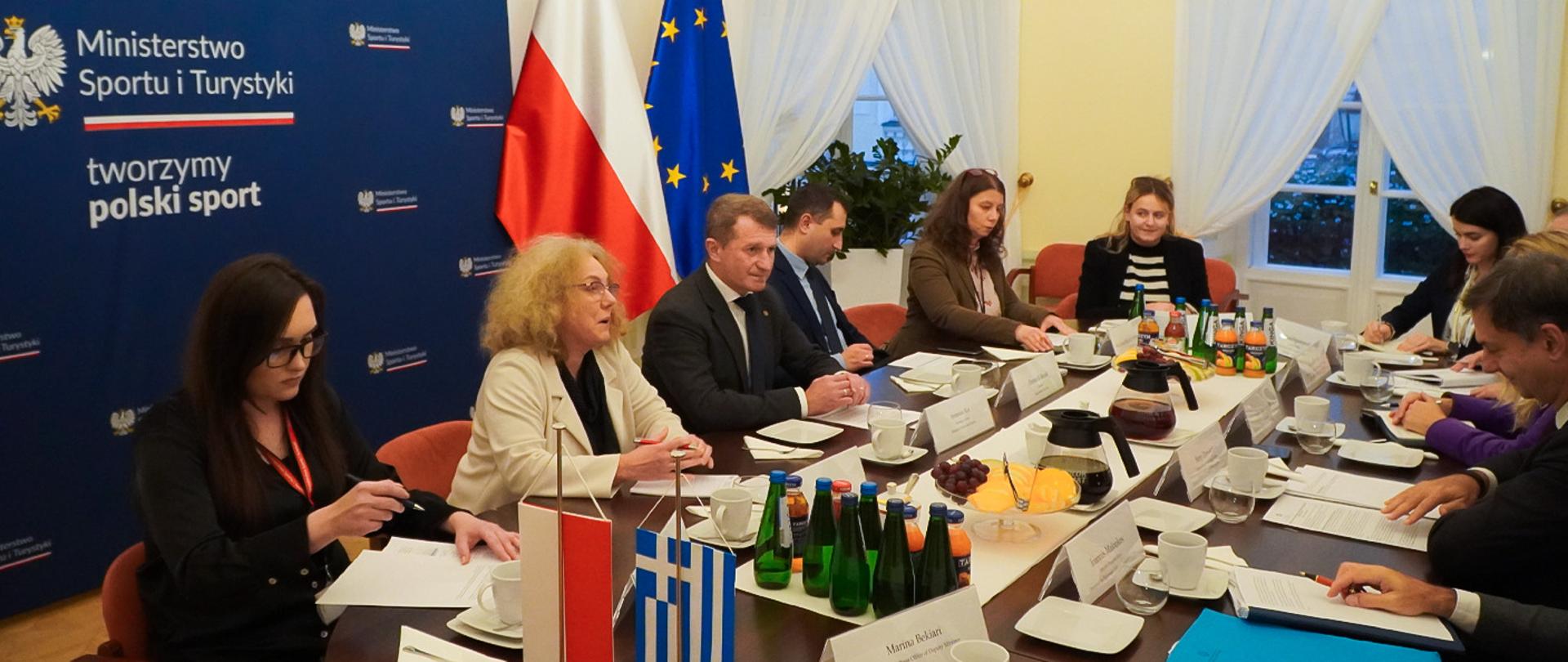 Spotkanie sekretarza stanu Ireneusza Rasia z wiceministrem spraw zagranicznych Grecji Harrym Theoharisem. Zdjęcie przedstawia ludzi siedzących przy stole. Na stole stoją napoje i owoce. Każdy uczestnik spotkania ma przed sobą kartki z wydrukami. Na stole stoją małe flagi Polski i Grecji. Za plecami uczestników spotkania granatowa ścianka z logotypem Ministerstwa Sportu i Turystyki oraz flagi Polski i UE.