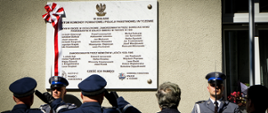 Przed tablicą przed którą stoi warta honorowa policjantów stoją policjanci oraz zaproszeni goście. Policjanci oddają honory.