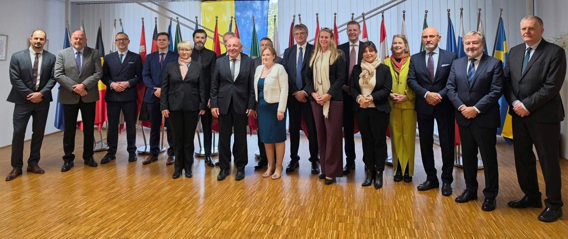 Zdjęcie grupowe uczestników spotkania dyplomatycznego. Piętnaście osób w formalnych strojach stoi w jednym rzędzie w sali konferencyjnej, na tle flag państw europejskich. Przed nimi drewniana podłoga, nad nimi jasny sufit z panelowym oświetleniem.