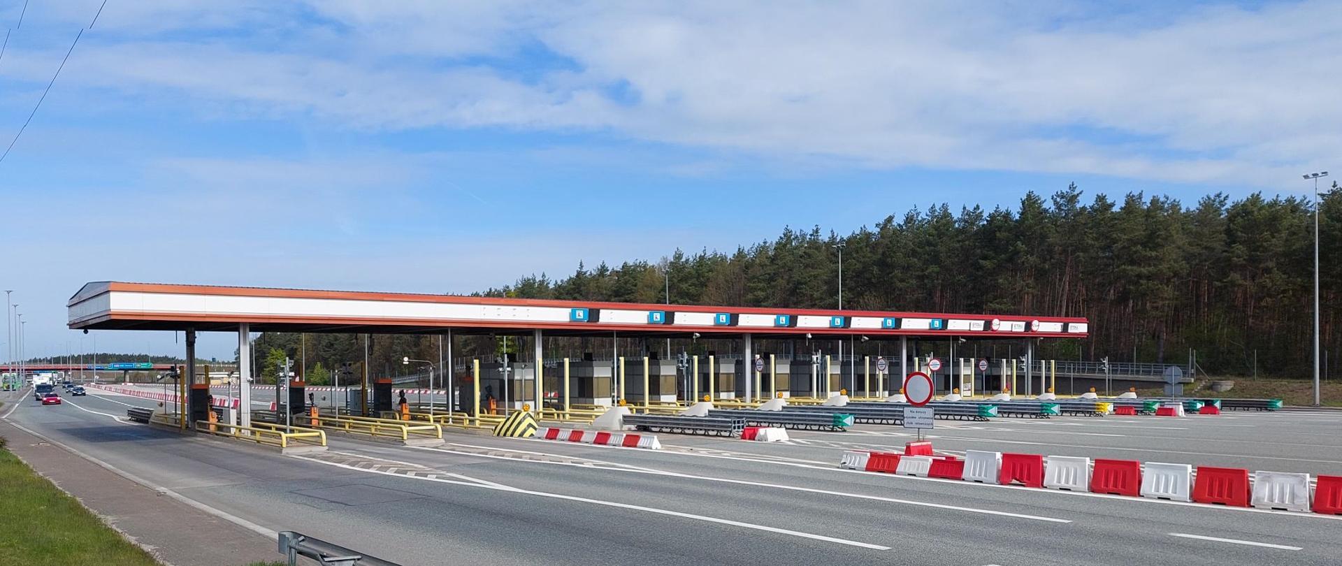 Punkt poboru opłat na autostradzie z wieloma pasami ruchu i zadaszonymi bramkami; przed nimi barierki i separatory w kolorach czerwonym, białym i zielonym. W tle widoczny las oraz pochmurne, jasnoniebieskie niebo, a po lewej stronie fragment drogi z nadjeżdżającymi pojazdami.