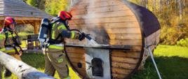 Zdjęcie przedstawia dwóch strażaków PSP, jeden leje wodę na saunę z której wydobywa się dym. 
