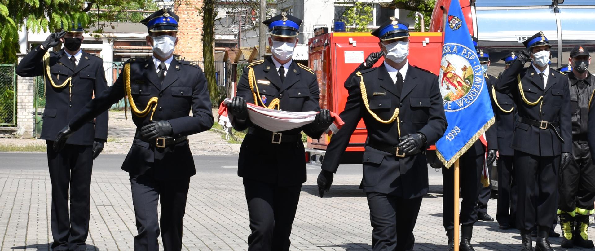 Na zdjęciu prezentuje się poczet flagowy KM PSP we Włocławku w trakcie uroczystego apelu. Umundurowanie funkcjonariuszy zgodne z ceremoniałem. Poczet podąża do miejsca podniesienia flagi.