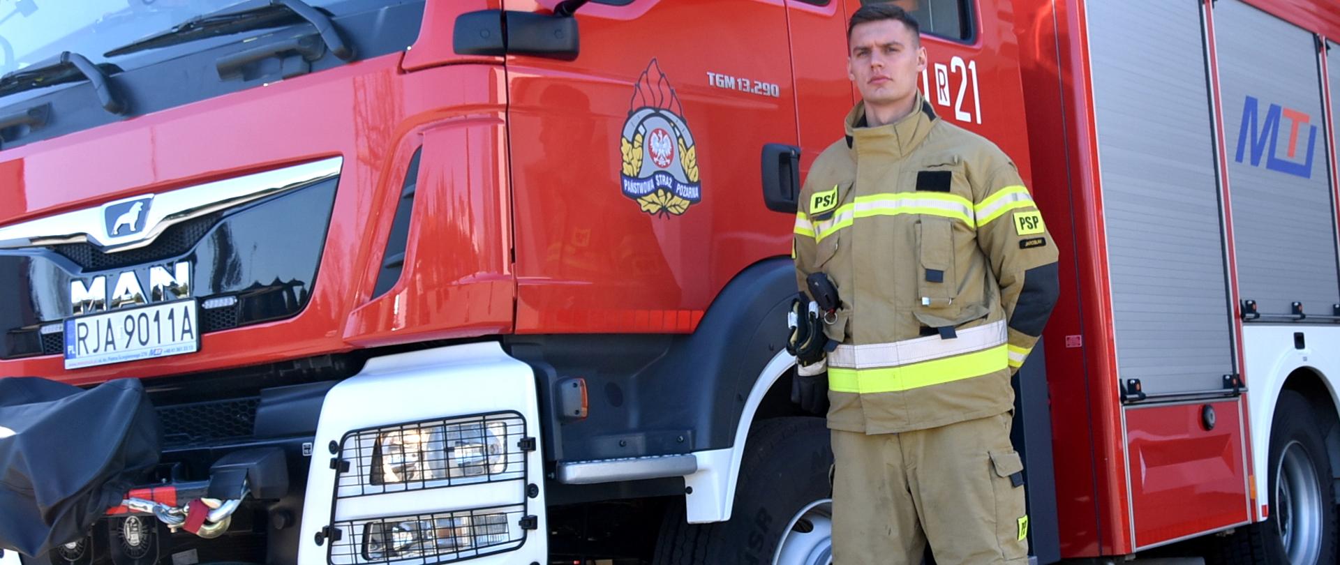 Na zdjęciu widać strażaka w umundurowaniu specjalnym, z rękami skrzyżowanymi za plecami. Za nim widać średni samochód ratowniczo-gaśniczy.
