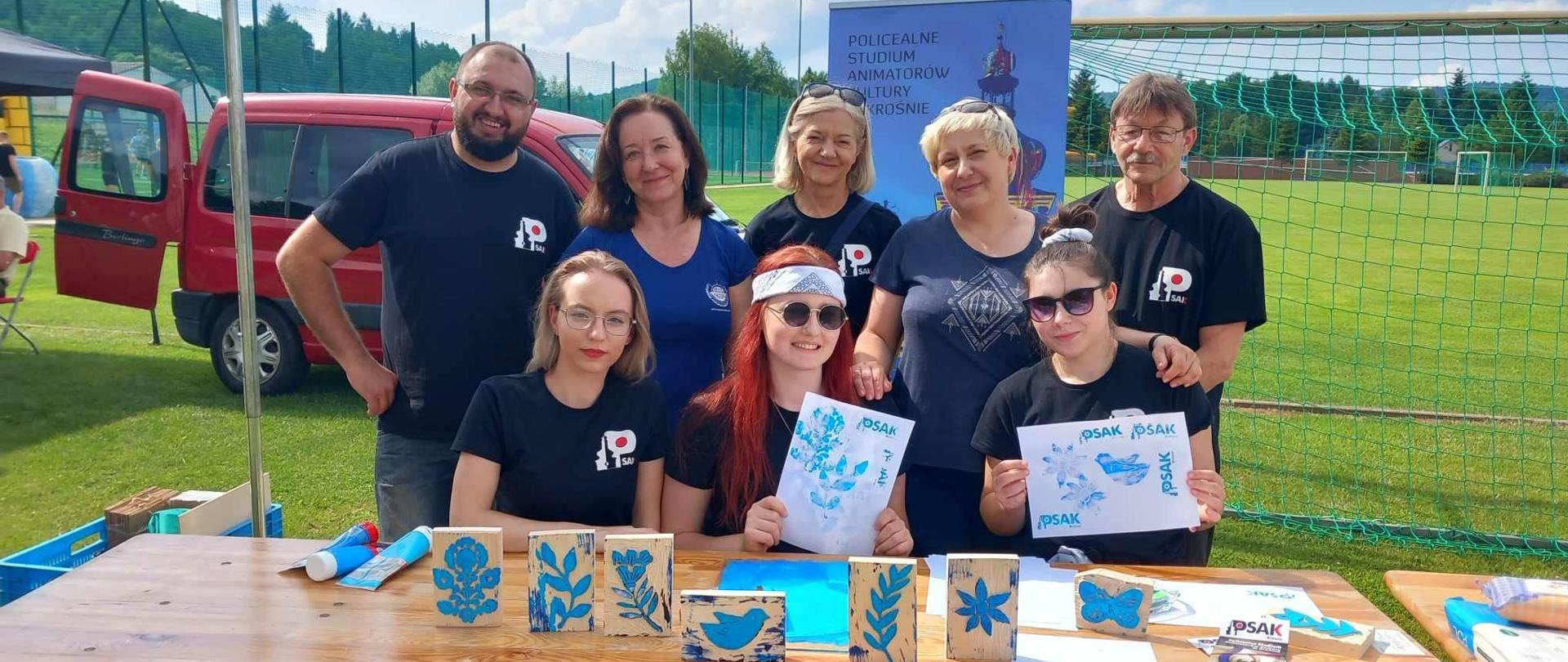 Słuchacze i nauczyciele podczas IV Festiwal Kultury Ludowej im. Stefana Kłosowicza. Przed nimi stoi stół z pracami plastycznymi. 