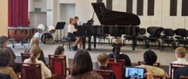 Dziewczynka i kobieta grają na fortepianie, z przodu widoczna publiczność.