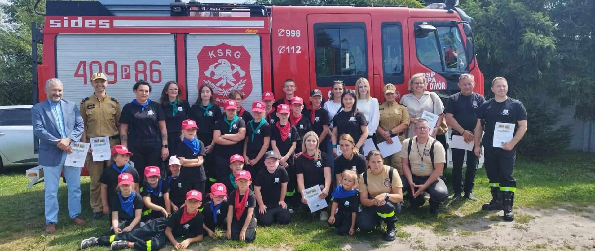 Zdjęcie przedstawia strażaków oraz dzieci i młodzież na wakacyjnym obozie strażackim.
W tle ośrodek wypoczynkowy.
