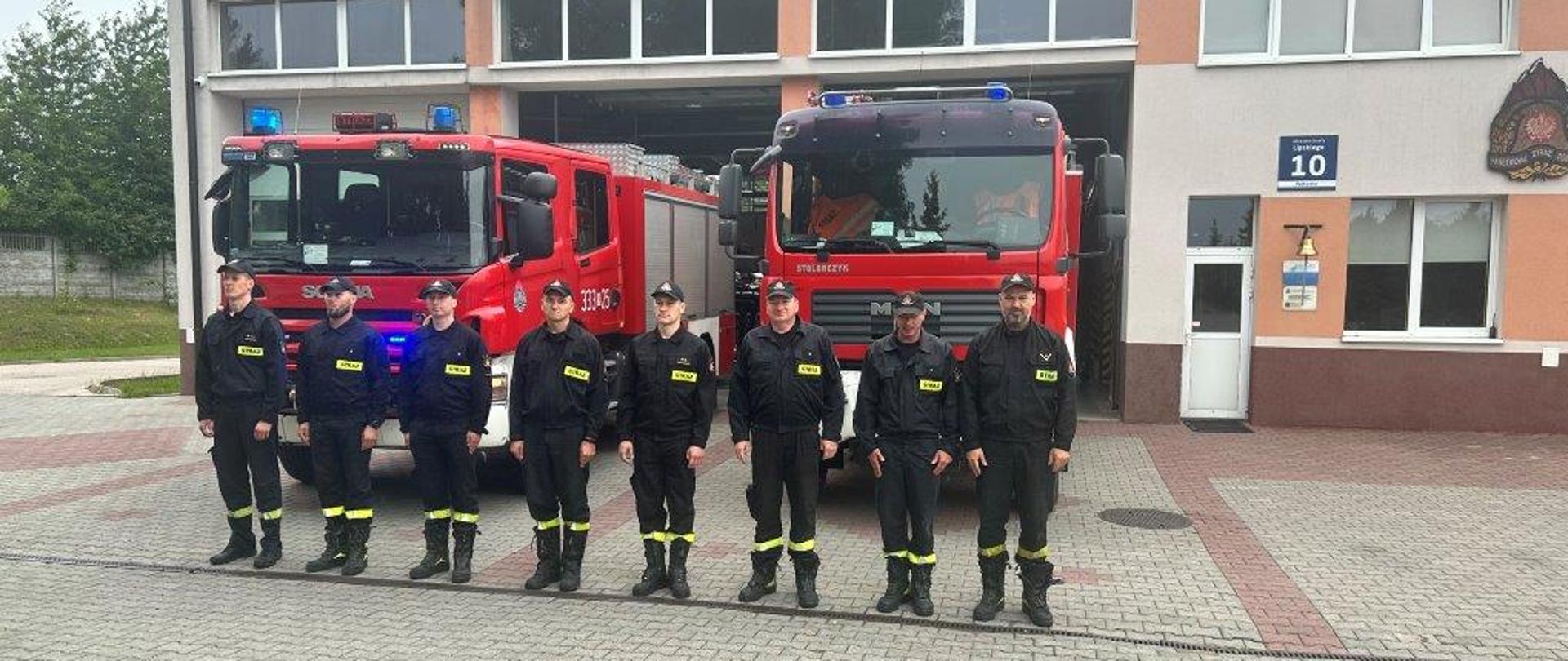 Zdjęcie wykonane przed jednostką ratowniczo – gaśniczą numer 3 w Radomiu. Na zdjęciu widać strażaków ustawionych w szeregu "na baczność" przed bramami garażowymi. Strażacy ubrani w ubrania koszarowe w kolorze czarnym z jaskrawymi elementami odblaskowymi. Na głowach mają czapeczki z daszkiem. Za strażakami znajdują się pojazdy ratowniczo - gaśnicze. Samochody w kolorze czerwonym z białymi oraz srebrnymi elementami zabudowy. Pojazdy mają zapalone niebieskie światła alarmowe oraz włączone sygnały dźwiękowe. Jeden ze strażaków - dowódca oddaje honory w celu upamiętnienia poległych w Powstaniu Warszawskim.