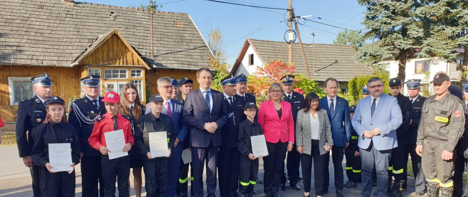 Zdjęcie przedstawia strażaków z Państwowej i Ochotniczej Straży Pożarnej w mundurach wyjściowych, kobiety i mężczyzn ubranych w ubrania cywilne, dzieci ubrane w mundury młodzieżowych drużyn pożarniczych OSP. W tle znajdują się domy oraz słup drewniany energetyczny.