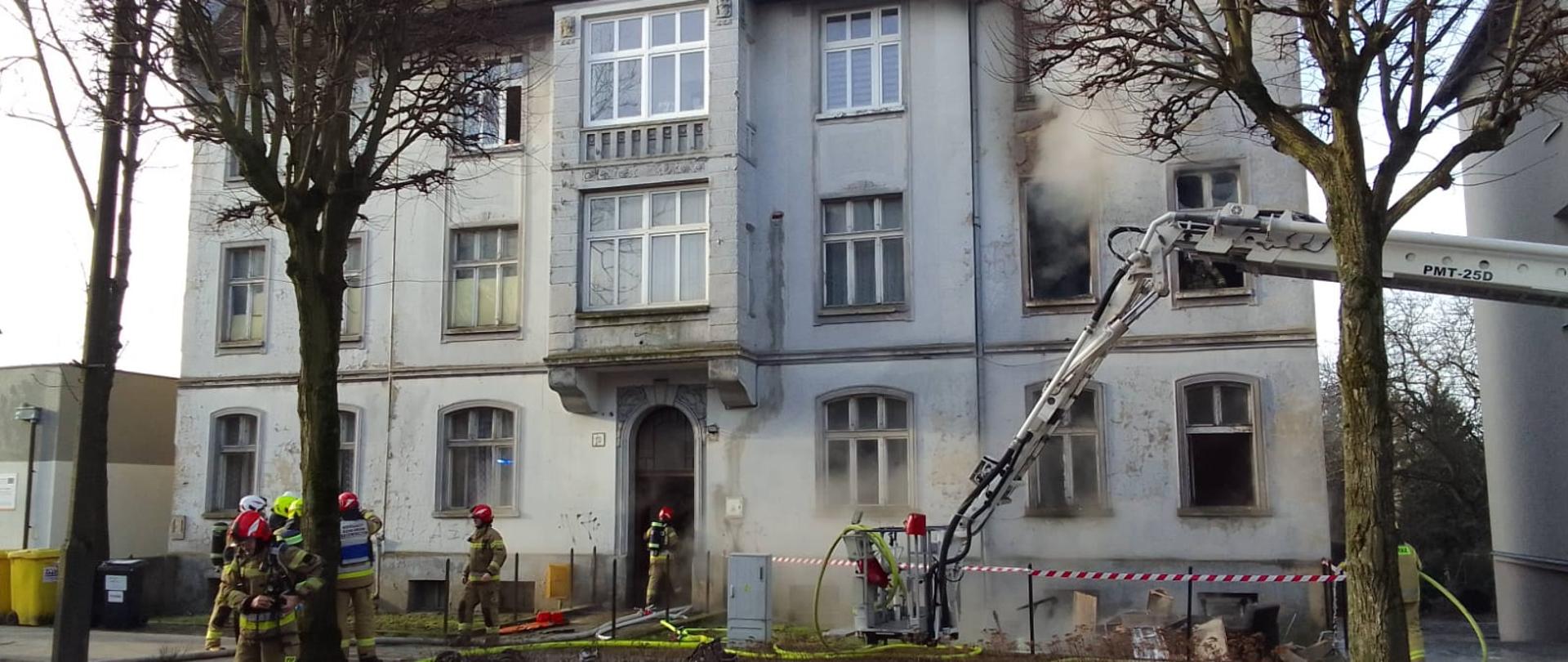 Dzień, na planie głównym budynek wielorodzinny kamienica z stylu z początku dwudziestego wieku z otworów okiennych na pierwszym piętrze z prawej strony widać wydobywający się siwy dym. Na tle budynku widać strażaków w trakcie prowadzenia akcji gaśniczej, ramie i kosz strażackiego podnośnika hydraulicznego oraz porozciągane węże. 