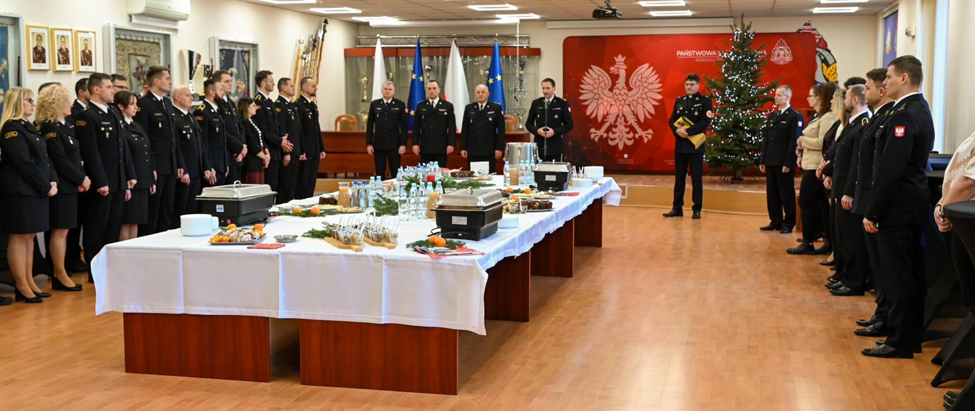 Szerokie ujęcie sali konferencyjnej. Uczestnicy spotkania opłatkowego stoją w dwóch rzędach wzdłuż długiego stołu z poczęstunkiem. Na końcu sali znajduje się choinka i oficjalne symbole państwowe.