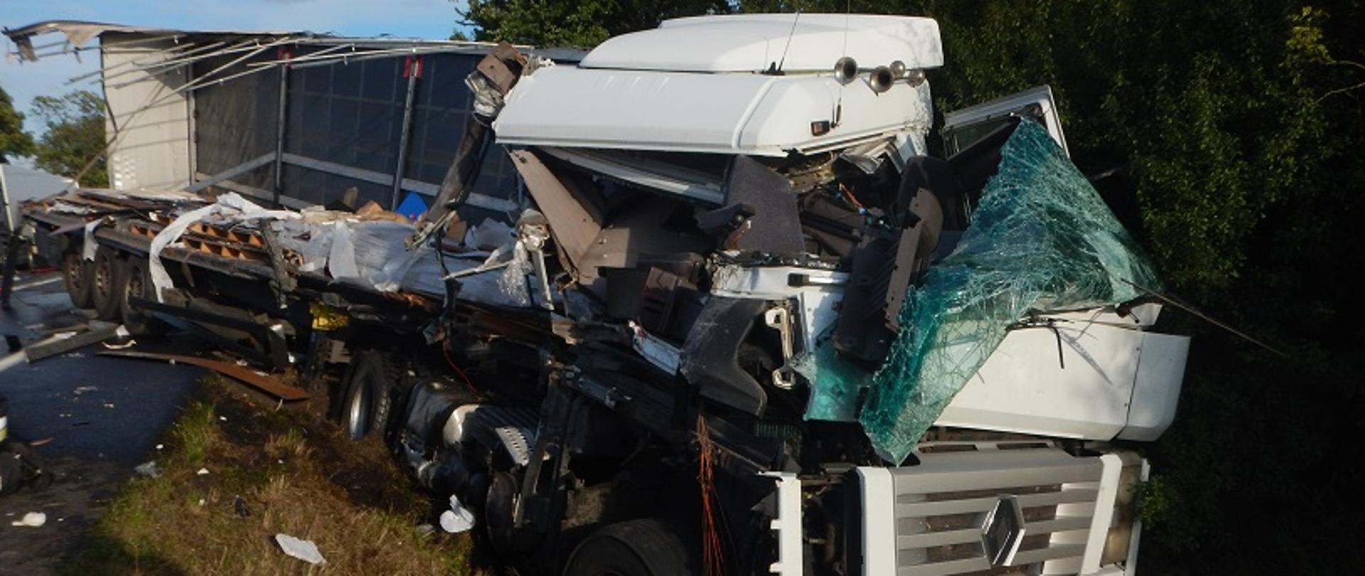 Zdjęcie przedstawia samochód ciężarowy, uszkodzony z prawej strony, który po zderzenie z innym samochodem ciężarowym znajduje się w rowie.