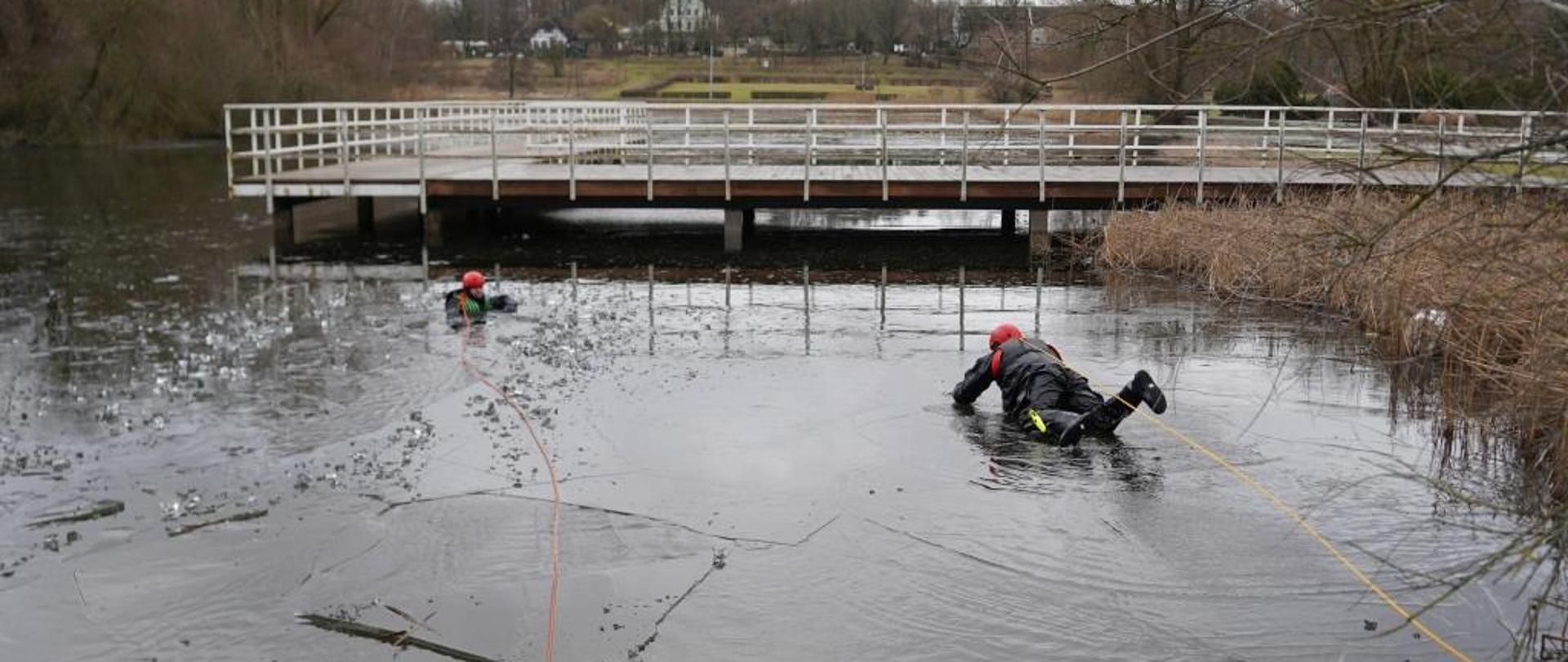 Po prawej stronie strażak w czarnym ubraniu wypornościowym czołga się po lodzie w kierunku poszkodowanego w przeręblu. Strażak zabezpieczony żółtą linką, osoba odgrywająca poszkodowanego zabezpieczona linką pomarańczową. 