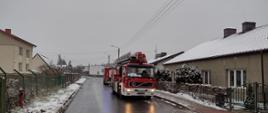 Na zdjęciu droga publiczna. Przy drodze budynki mieszkalne. Na budynkach śnieg. Zachmurzenie duże. Pora zimowa. Na drodze stoją auta ratowniczo-gaśnicze w kolorze czerwonym z sygnalizacją ostrzegawczą.