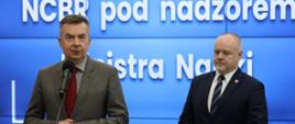Na tle wbudowanego w ścianę wielkiego ekranu z napisem NCBR pod nadzorem Ministra Nauki stoi minister Wieczorek, który mówi do mikrofonu na stojaku, obok niego wiceminister Gzik.