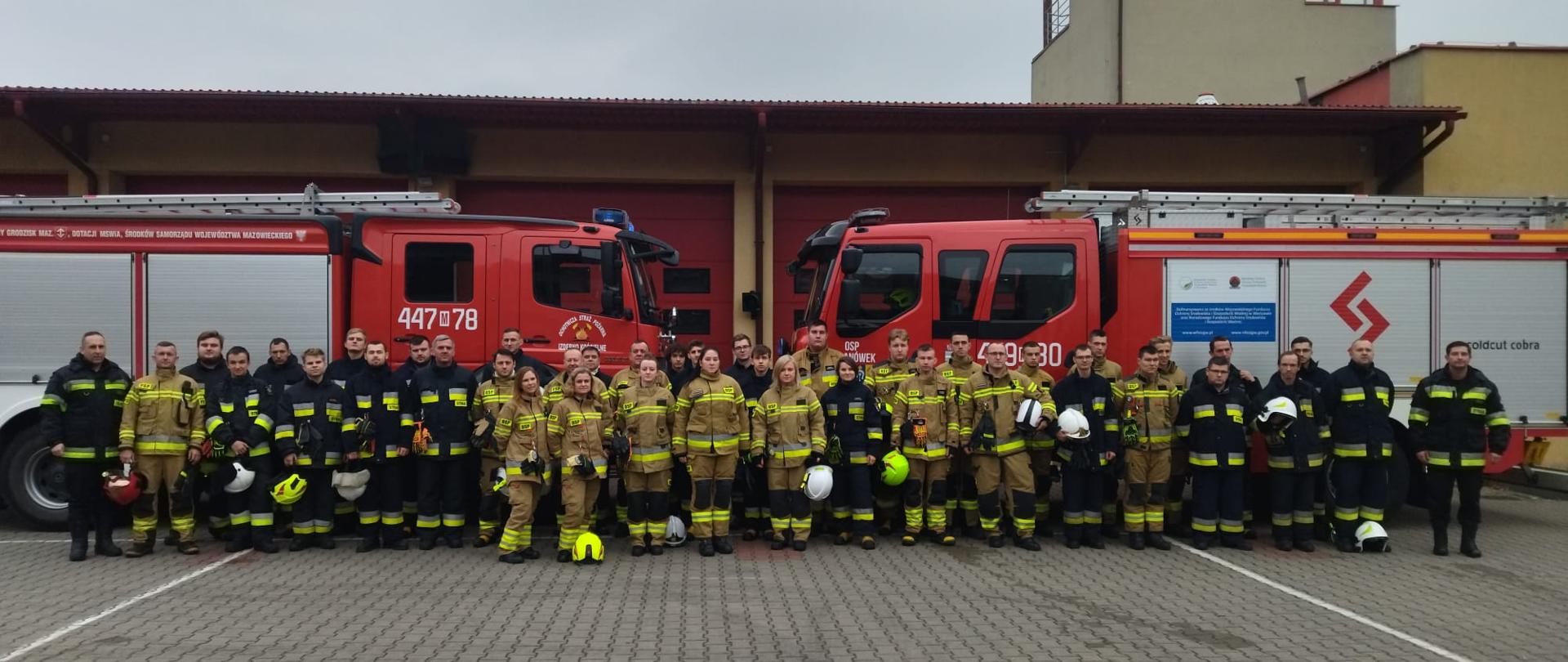 Zdjęcie grupowe strażaków ochotników i komisji egzaminacyjnej uczestniczących w egzaminie kończącym „Szkolenie podstawowe strażaka ratownika OSP” na tle samochodów pożarniczych oraz bram garażowych strażnicy JRG Grodzisk Maz.