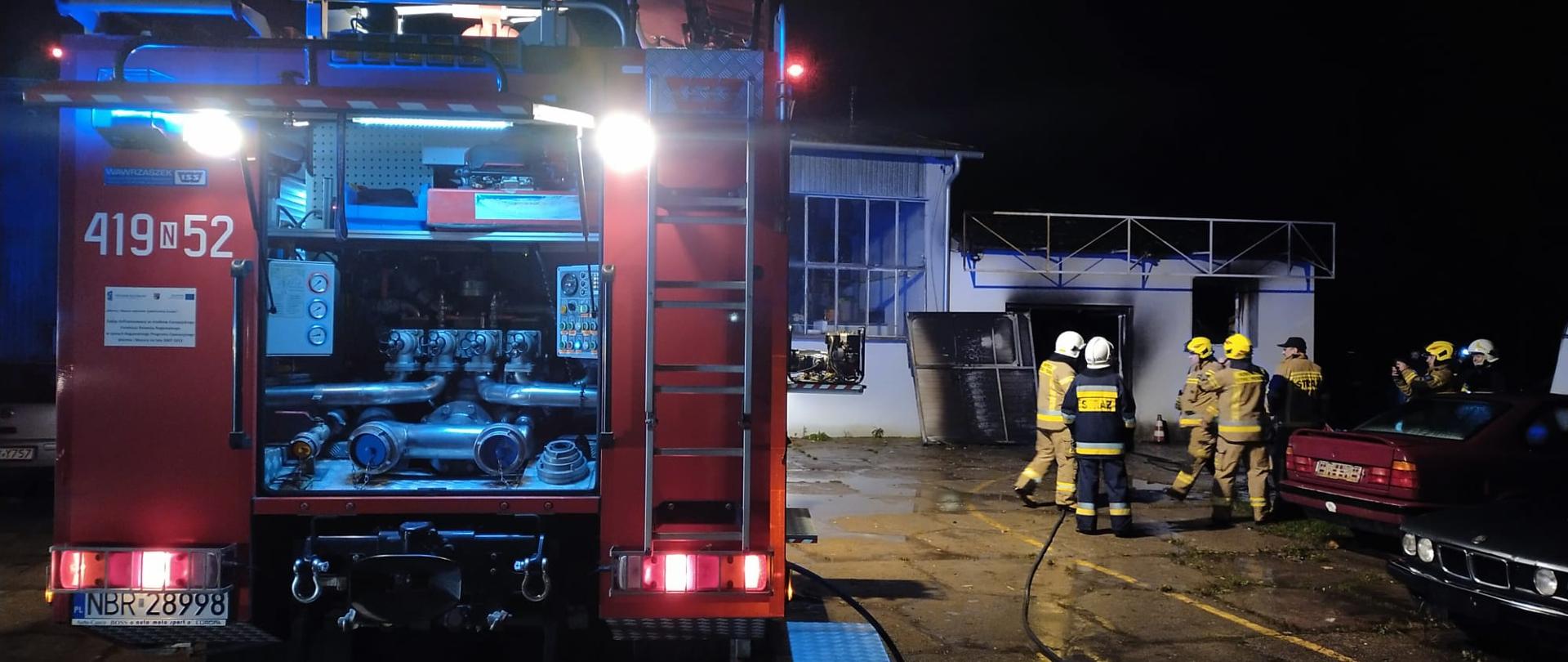 Zdjęcie zrobione nocą. Z lewej strony oświetlony tył pojazdu strażackiego, otwarta skrytka samochodu. Z prawej strony strażacy i samochody osobowe. W oddali biały budynek po pożarze.