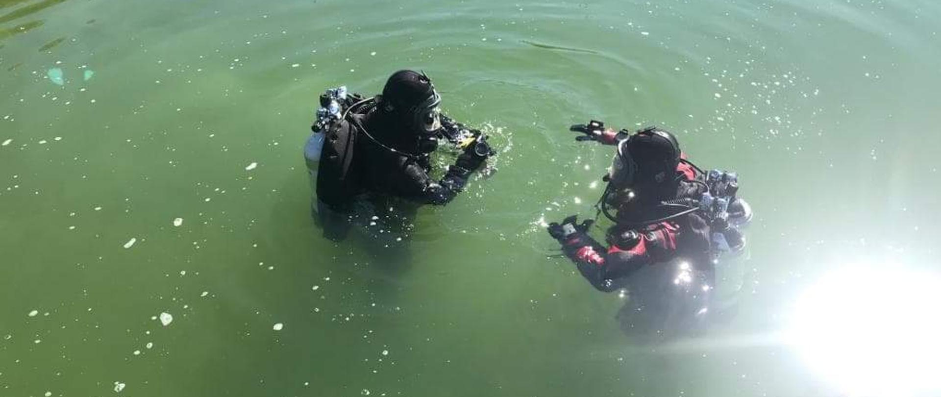 Dwóch nurków stoi w wodzie do pasa i przygotowuje się do zejścia pod wodę. 