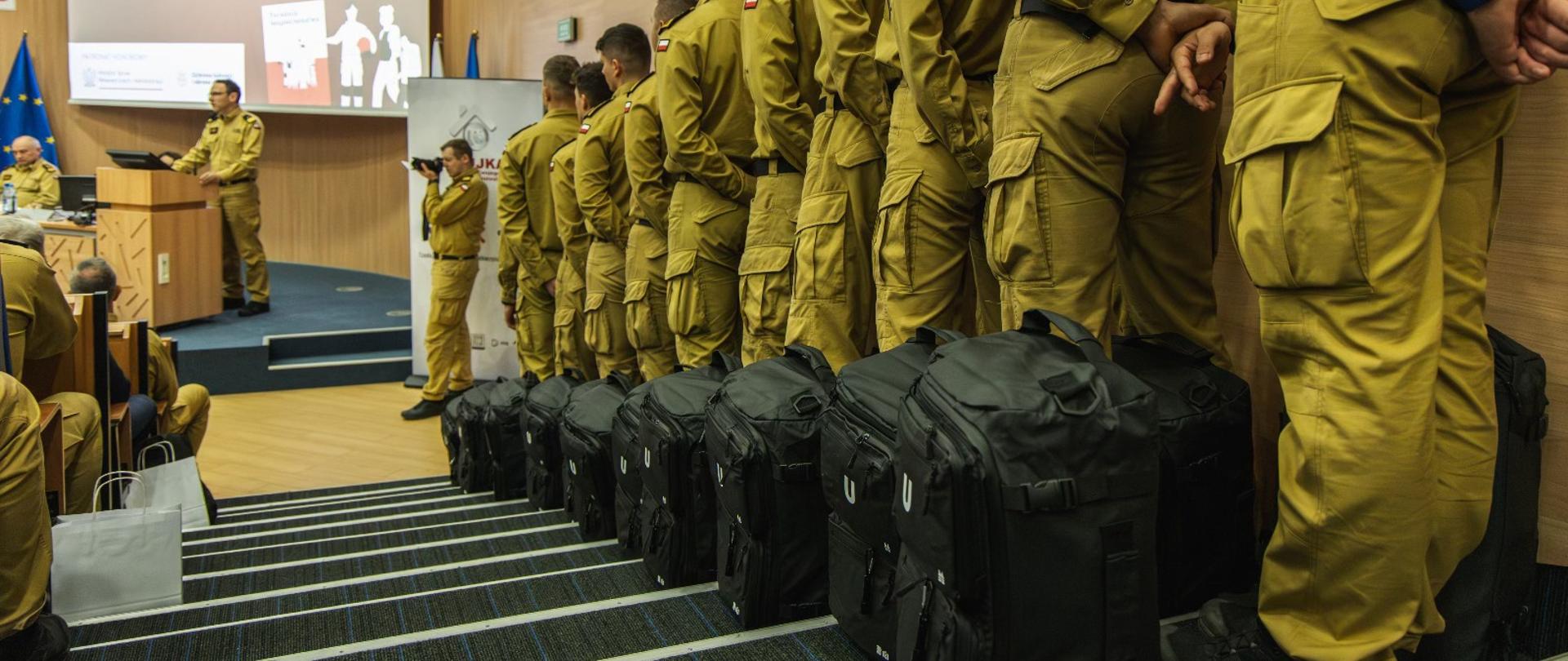 duża aula, w szeregu na schodach stoją strażacy, przy nich duże czarne plecaki 