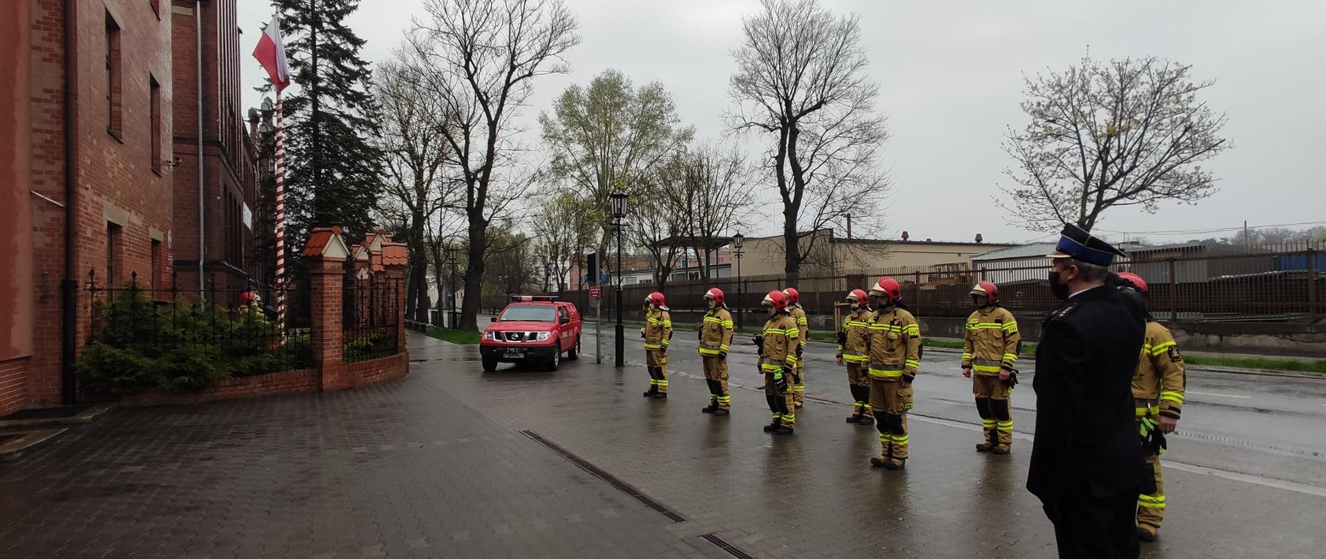 Na zdjęciu widać całą zmianę służbową, która uczestniczy w podniesieniu flagi państwowej na maszt. Strażacy ustawieni są w dwuszeregu. Na wysokości masztu znajduje się samochód strażacki
