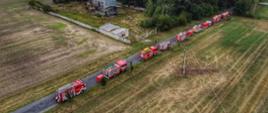 Widok z drona na dziewięć zastępów wozów strażackich stojących za sobą i przegotowanych do rozpoczęcia ćwiczeń.