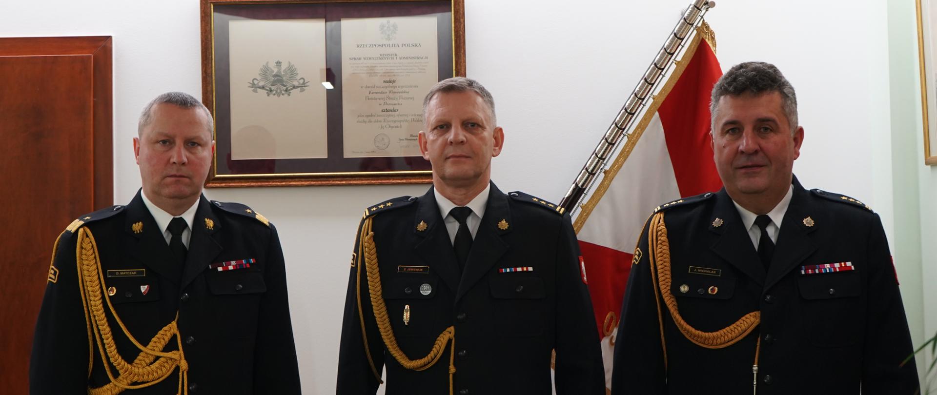trzech strażaków w ciemnych mundurach ze sznurem stoi na baczność obok siebie. w tle sztandar
