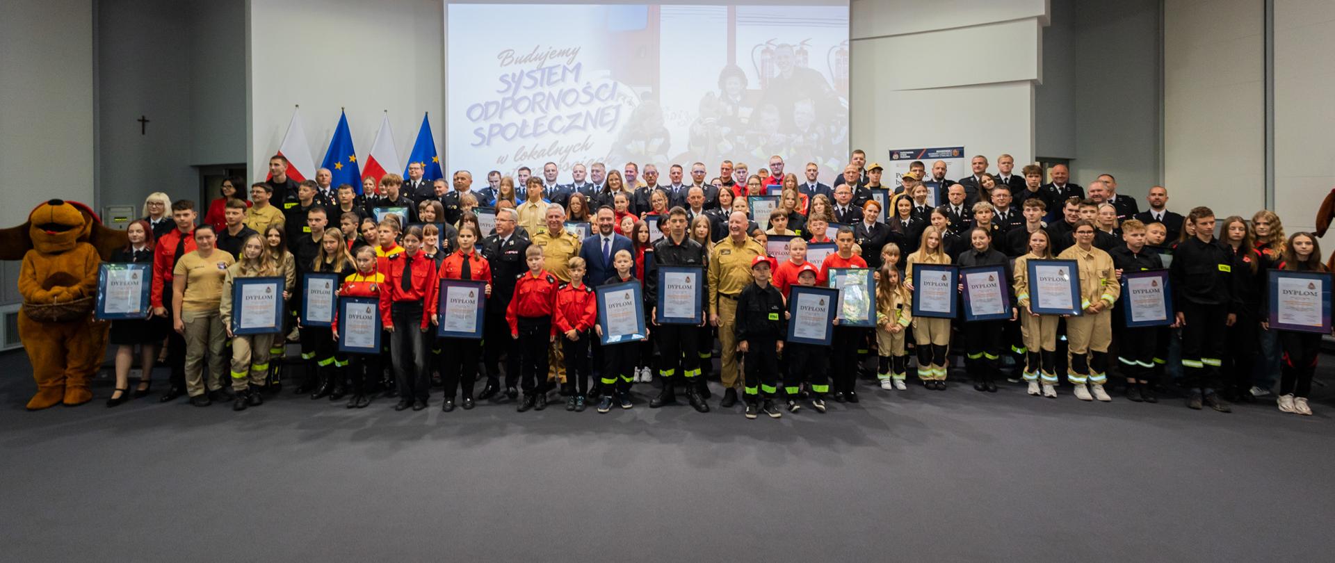 Bardzo duża grupa uczestników, w tym dzieci i młodzież w mundurach strażackich, stoi na scenie w obszernej sali konferencyjnej. Wielu laureatów trzyma w rękach oprawione w czarne ramki dyplomy uznania. W tle na ekranie wyświetla się napis „Budujemy SYSTEM ODPORNOŚCI SPOŁECZNEJ w lokalnych społecznościach”, a obok wiszą flagi Polski i Unii Europejskiej. 