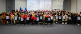 Bardzo duża grupa uczestników, w tym dzieci i młodzież w mundurach strażackich, stoi na scenie w obszernej sali konferencyjnej. Wielu laureatów trzyma w rękach oprawione w czarne ramki dyplomy uznania. W tle na ekranie wyświetla się napis „Budujemy SYSTEM ODPORNOŚCI SPOŁECZNEJ w lokalnych społecznościach”, a obok wiszą flagi Polski i Unii Europejskiej. 