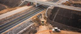 Widok z lotu ptaka na budowę węzła drogowego: nowy wiadukt nad drogą ekspresową, oznakowane jezdnie, skarpy drogowe oraz pracujący sprzęt budowlany i pojazdy techniczne.