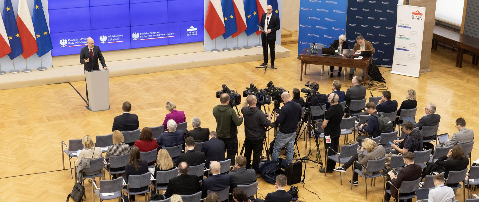 Minister, członek Rady Ministrów Polski Maciej Berek przemawia zza pulpitu mównicy. W tle widoczny ekran, na którym wyświetlana jest nazwa Wydarzenia oraz stojące po obu stronach ekranu flagi Polski i Unii Europejskiej, a na pierwszym planie rzędy krzeseł, na których zasiada publiczność - uczestnicy wysłuchania obywatelskiego.