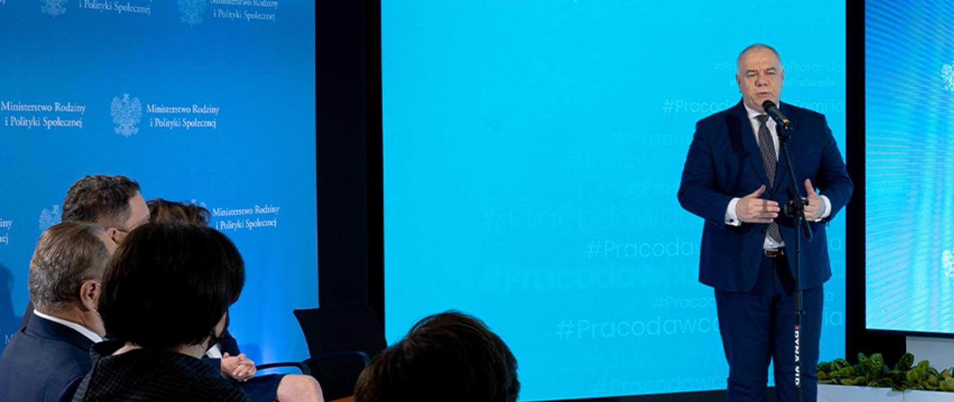 Wicepremier Jacek Sasin stoi przy mikrofonie. Po lewej stronie uczestnicy debaty. W tle telebim z logotypami MAP i MRiPS.